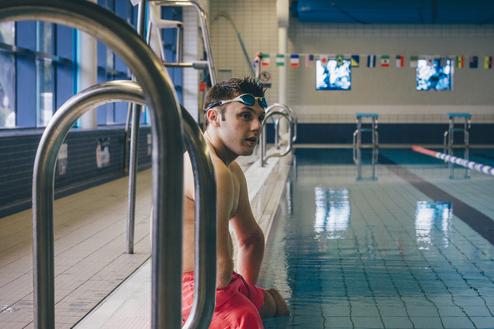Mann in Badeshorts sitzt am Beckenrand eines Schwimmbads, Blick zum Wasser, Schwimmbrille auf dem Kopf.