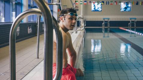Mann in Badeshorts sitzt am Beckenrand eines Schwimmbads, Blick zum Wasser, Schwimmbrille auf dem Kopf.