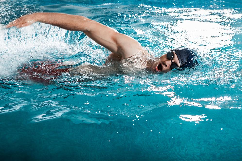 Mann schwimmt im Freistilstil in einem hellblauen Schwimmbecken, Spritzer Wasser sind um ihn herum zu sehen.