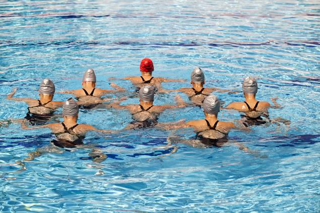 Gruppe von Synchron-Schwimmerinnen in Badeanzügen und Badekappen, die in einem Schwimmbecken synchron schwimmen.
