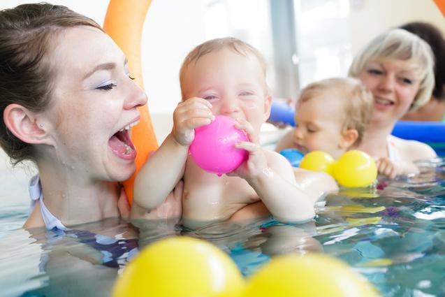 Mutter und Kleinkind im Wasser, wobei das Kind einen pinken Ball hält und beide lachen. Bunte Wasserspielzeuge um sie herum.