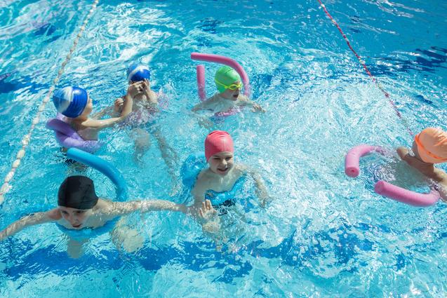 Sechs Kinder schwimmen in einem hellblauen Pool, einige mit Schwimmnudeln und farbigen Badekappen.