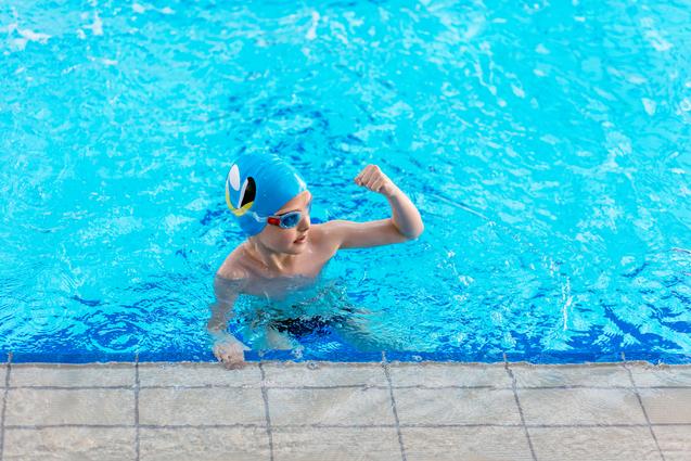 Junge mit blauer Schwimmhaube und Schwimmbrille zeigt seinen Muskel im Wasser eines Schwimmbeckens.