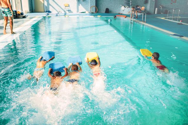 Fünf Kinder schwimmen im Becken und nutzen Schwimmbretter in einem hell erleuchteten Hallenbad.