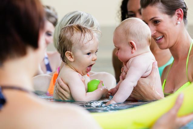 Zwei Babys im Wasser: eines mit einem grünen Ball, das andere in den Händen einer Frau. Wasser spritzt um sie herum.