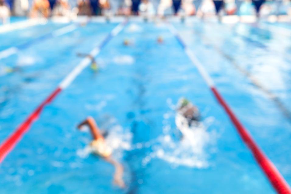 Unschärfe eines Schwimmwettkampfs in einem Becken mit Schwimmern und Zuschauern am Beckenrand.