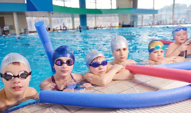 Sechs Kinder im Schwimmbecken lehnen mit Schwimmbrillen und -kappen an der Beckenkante, Schwimmnudeln neben ihnen.