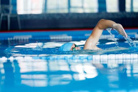 Eine Person mit blauer Badekappe und Schwimmbrille schwimmt in einem Hallenbad und spritzt Wasser mit der Hand.