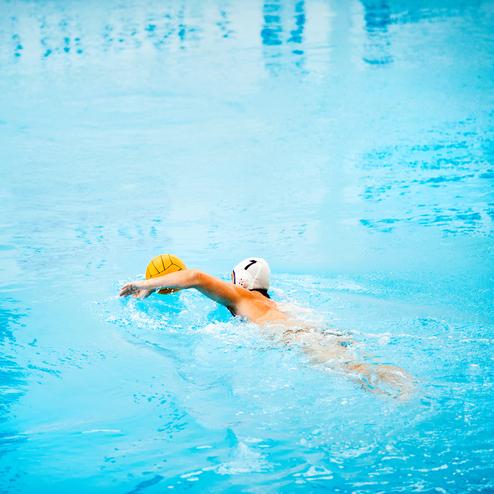 Ein Schwimmer mit einer weißen Badekappe verfolgt einen Wasserball in einem blauen Schwimmbecken.