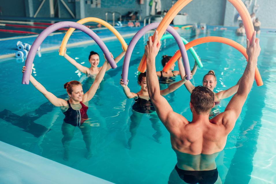 Gruppen von Schwimmern heben bunte Poolnudeln im Wasser während eines Aquatrainings in einem Hallenbad.